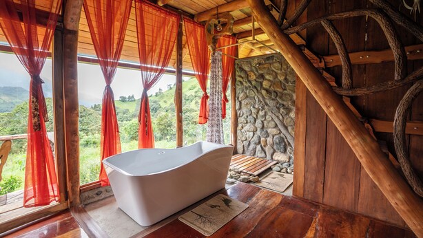 A rustic bathroom with a freestanding bathtub, wooden beams, and a stone wall. - Amanecer Andino (Rest cabin in the mountain) (Puyo)