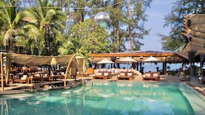 A pool area with lounge chairs and umbrellas, surrounded by palm trees and a thatched-roof structure. - Cafe del Mar Hotel (Kamala)