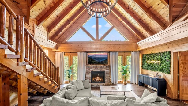 A spacious living room with a stone fireplace, wooden staircase, and a large sofa. - Luxury 7BR Smoky Mountain Lodge, Outdoor Pool, Hot Tub, Game Room & Theater -24 (Sevierville)