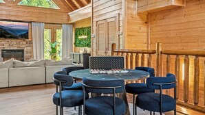 A wooden cabin interior with a round table, blue chairs, a sofa, a fireplace, and a balcony with a wooden railing. - Luxury 7BR Smoky Mountain Lodge, Outdoor Pool, Hot Tub, Game Room & Theater -24 (Sevierville)