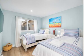 A bedroom with two beds, a window with curtains, a painting of a beach scene, and a bedside table with a lamp.