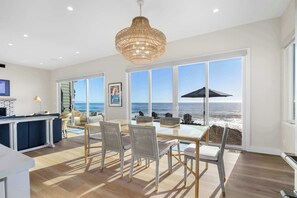 A spacious dining area with a view of the ocean, featuring a large table and chairs, a hanging light fixture, and a window with a view of the beach.
