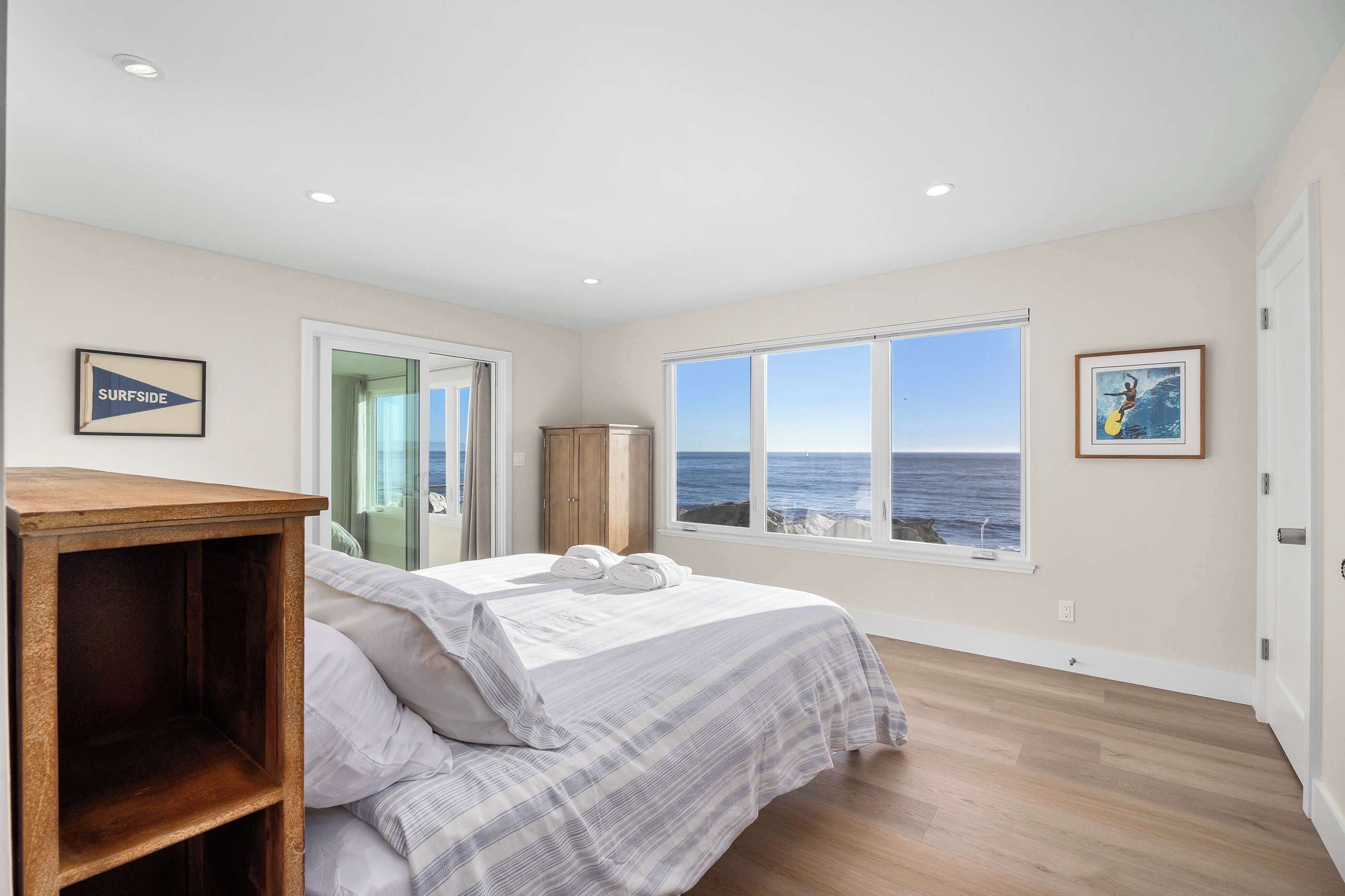 Un dormitorio con cama, mesita de noche de madera, armario y un amplio ventanal con vista al océano.