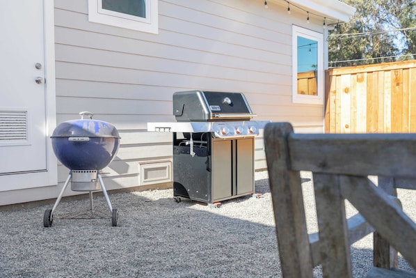 A portable grill set up on a gravel surface in front of a house with a wooden fence.
