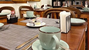 Zona pranzo con tavolo di legno apparecchiata per due persone, una grande finestra con stipiti in legno e una libreria piena di libri e oggetti decorativi.