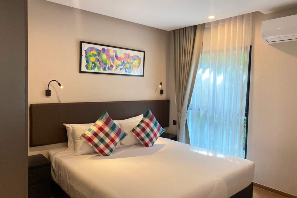 A hotel room with a large bed, two pillows, a framed artwork, a window with curtains, and an air conditioning unit.