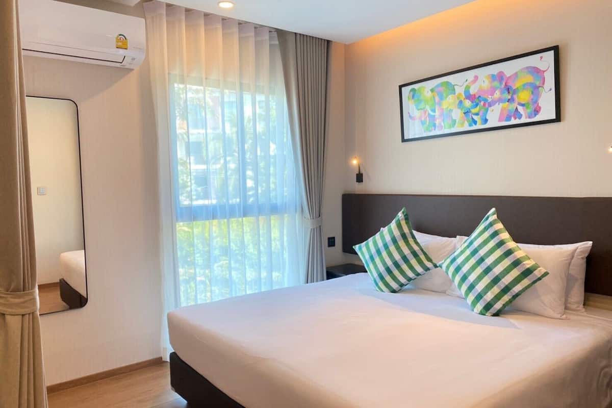 A hotel room with a large bed, a window with sheer curtains, a wall-mounted air conditioner, and a colorful abstract painting.