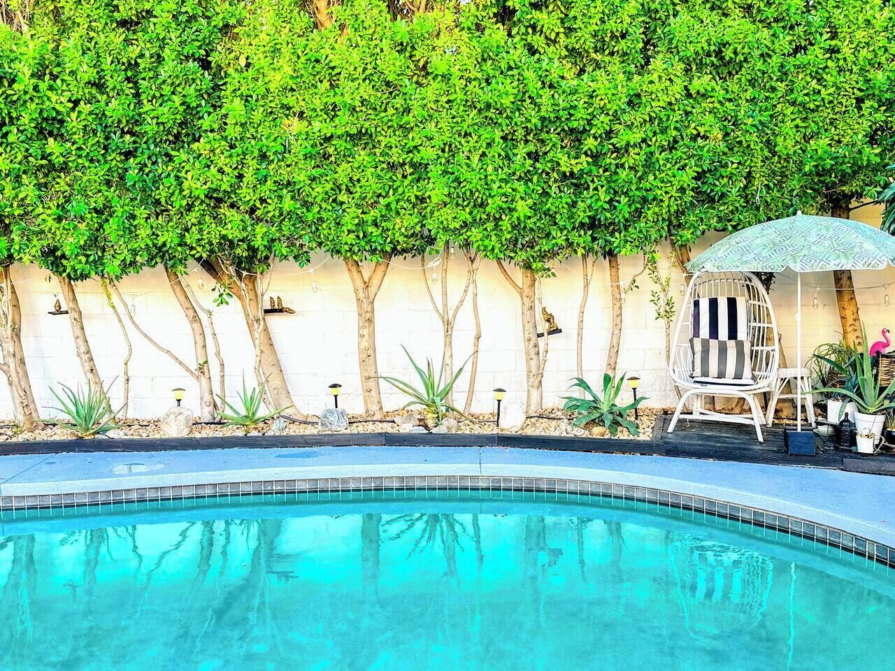A swimming pool with a tiled edge, surrounded by a low wall with trees and plants. There is a white chair with a striped cushion and a small umbrella.