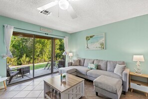 Ein Wohnzimmer mit einer Couch, einem Couchtisch und einer Lampe. Ein Schiebetürfenster führt auf eine Terrasse mit Tisch und Stühlen.
