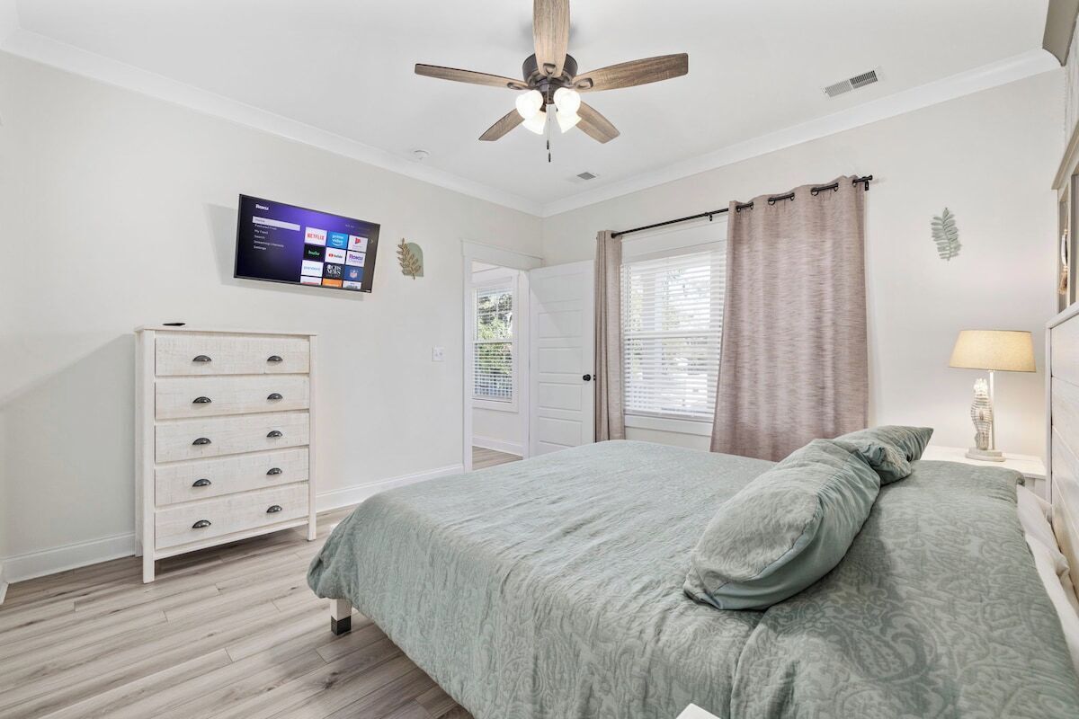 Une chambre à coucher comprenant un lit, un ventilateur de plafond, un téléviseur fixé au mur, une commode et une fenêtre avec des rideaux.