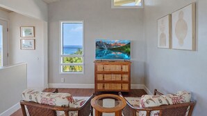 Ein Wohnzimmer mit einem großen Fernseher auf einer Holzkommode, einem Fenster mit Blick auf den Ozean und einem Sofa mit gemusterten Kissen.