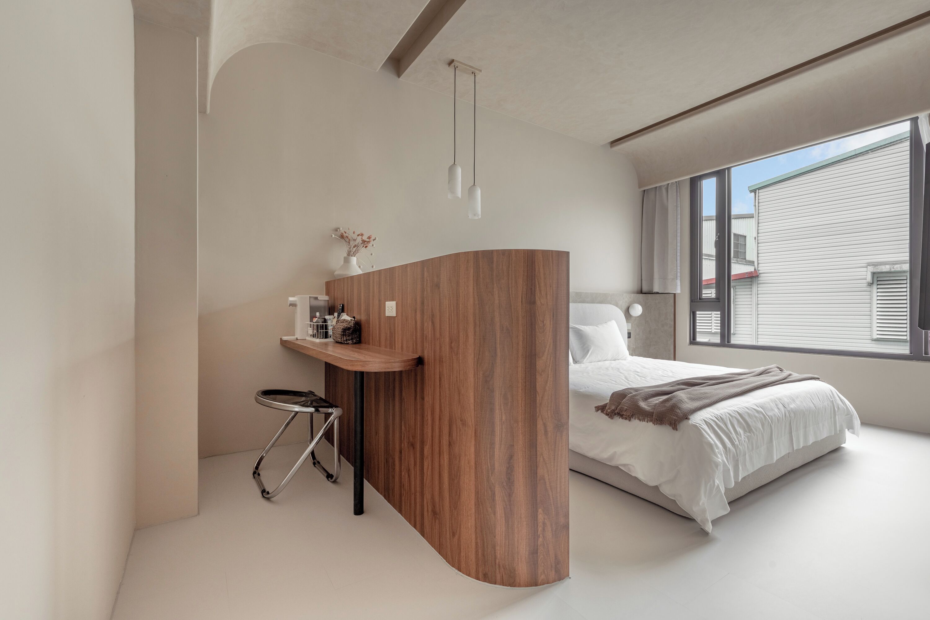 Un dormitorio moderno con un escritorio de madera curvada, cama con ropa de cama blanca y un gran ventanal que proporciona luz natural.