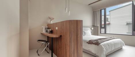 Un dormitorio moderno con un escritorio de madera curvada, cama con ropa de cama blanca y un gran ventanal que proporciona luz natural.