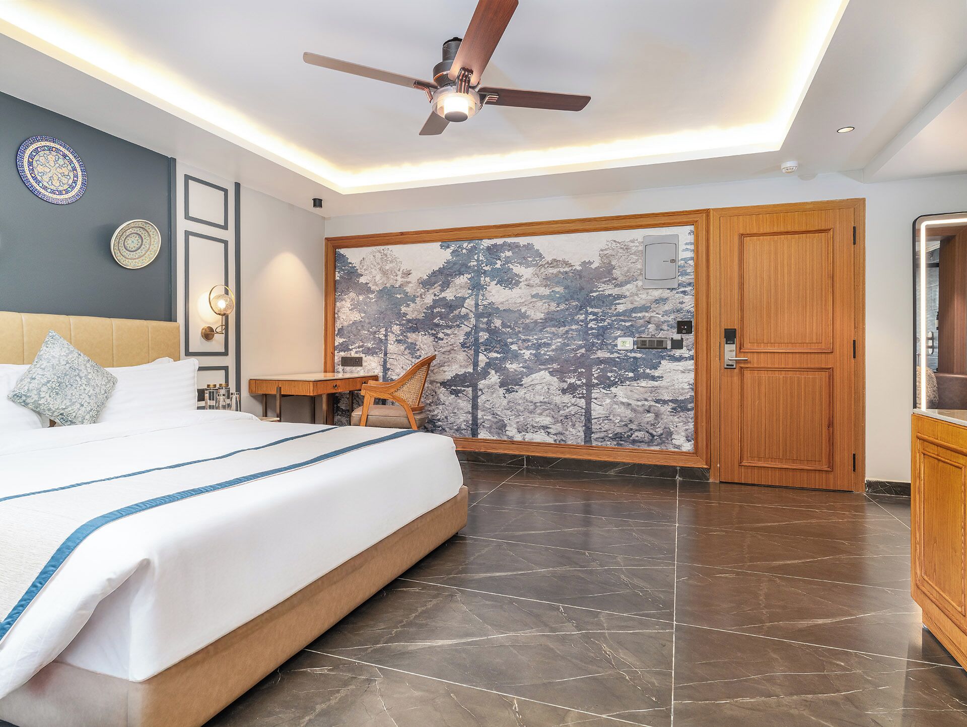 A modern hotel room with a large bed, a desk, a chair, a ceiling fan, and a large window with a forest mural.