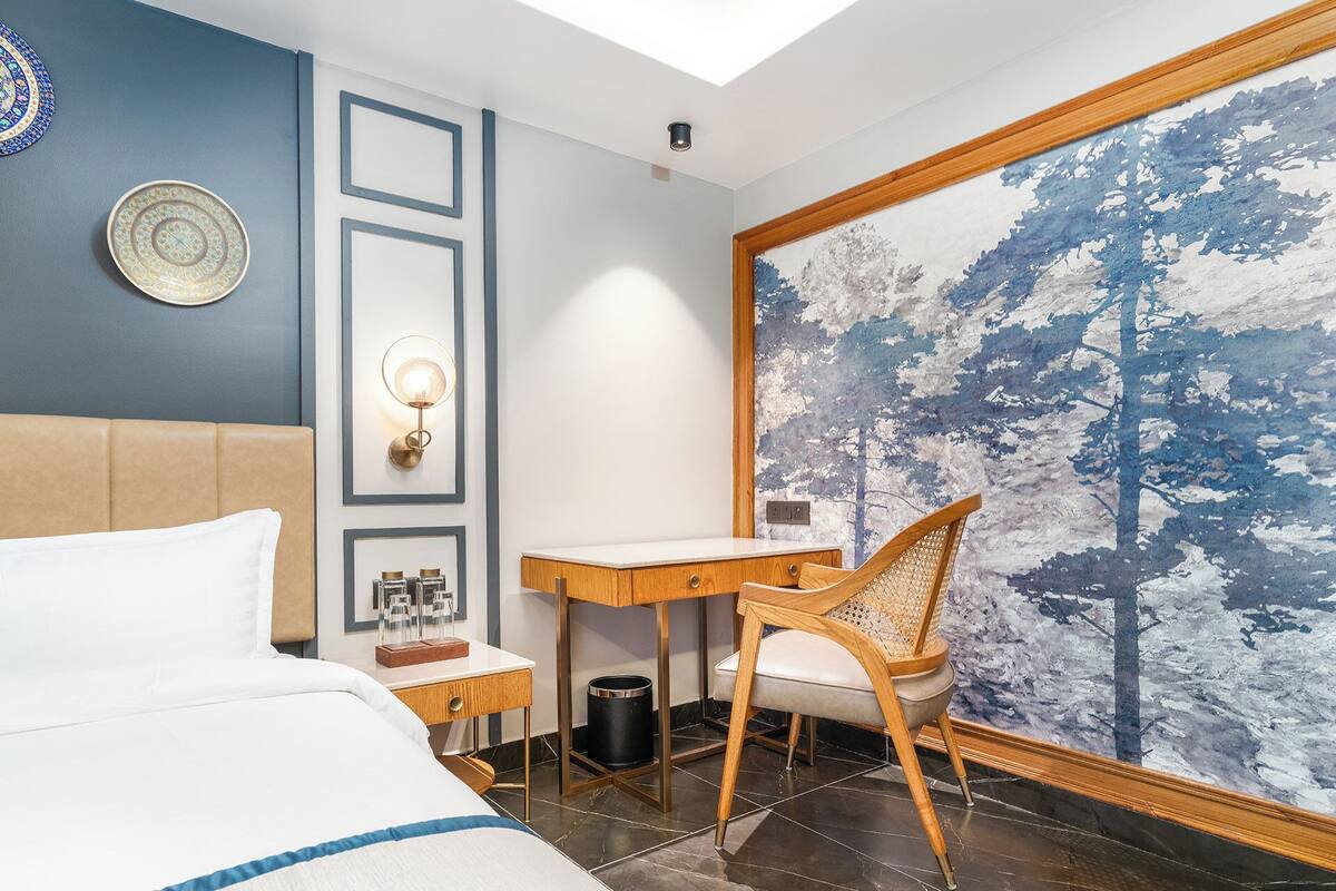 A hotel room with a bed, a desk, a chair, a nightstand, and a large window with a forest mural.