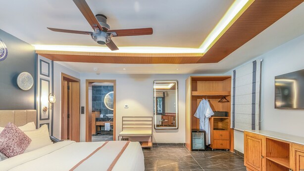 A modern hotel room with a large bed, a ceiling fan, a TV, and a walk-in closet. - The Vale Lodge (Anantnag)