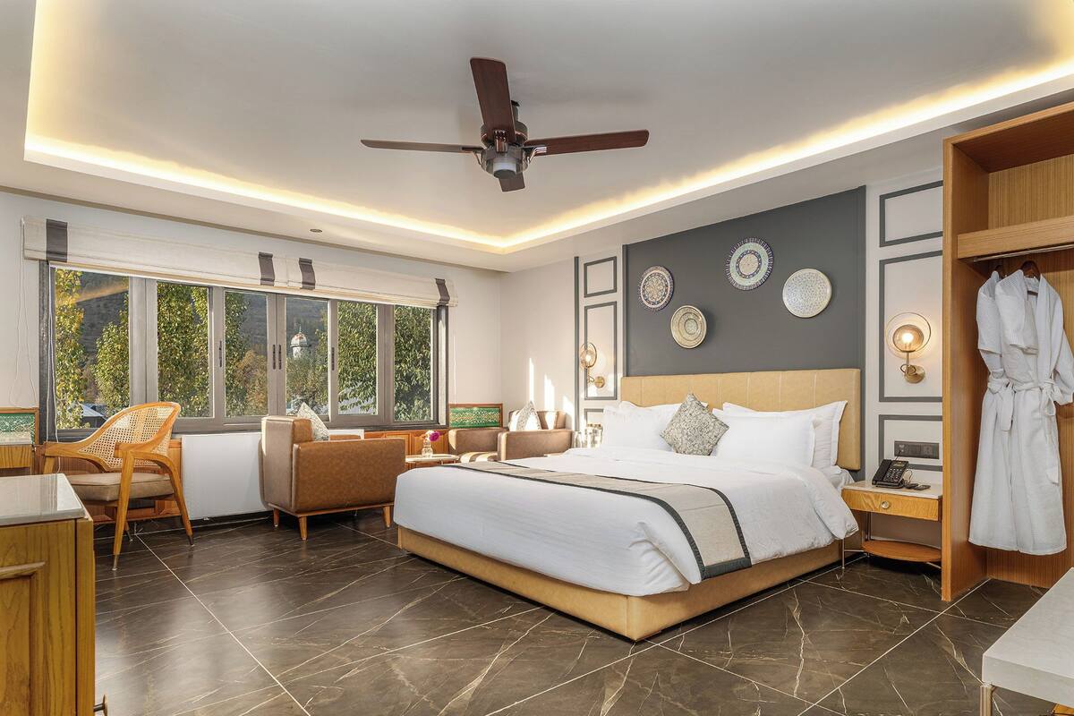 A modern hotel room with a large bed, a sitting area with a chair, a wardrobe, and a view of greenery through the windows.
