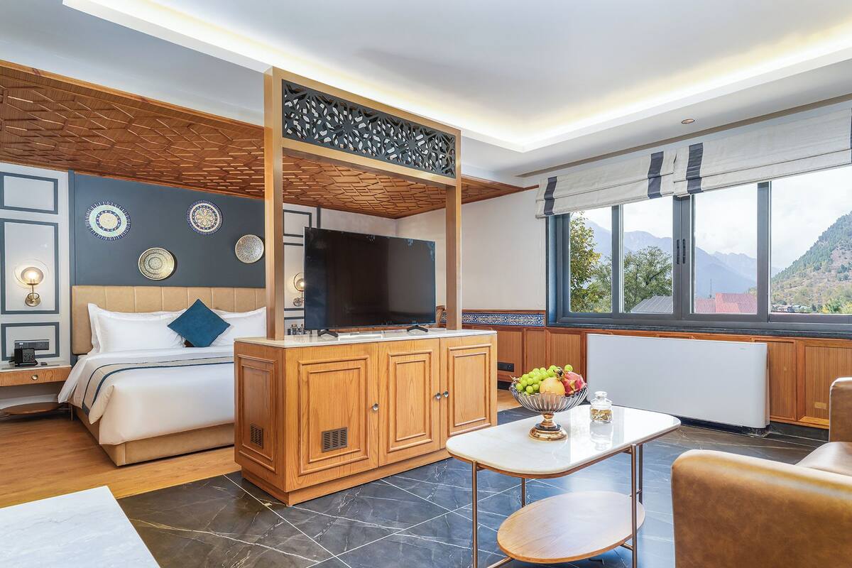 A modern hotel room with a bed, a TV, a wooden cabinet, a dining table, and a view of the mountains.