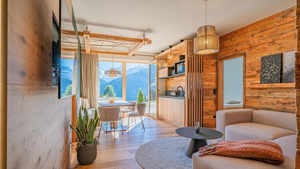 Un salon moderne avec des murs en bois, un canapé, un coin repas comprenant une table et des chaises, et une vue sur les montagnes grâce à de grandes fenêtres.