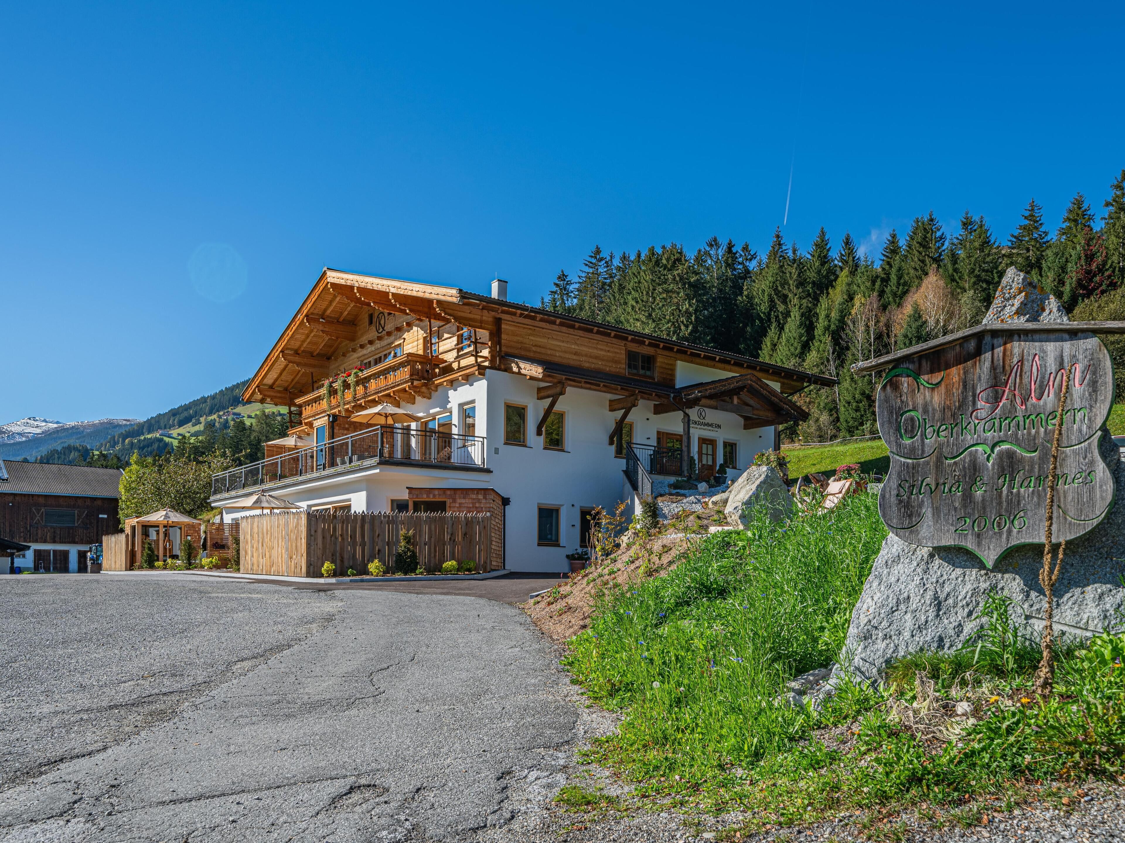 A two-story chalet with a wooden balcony, a stone sign, and a mountainous backdrop.