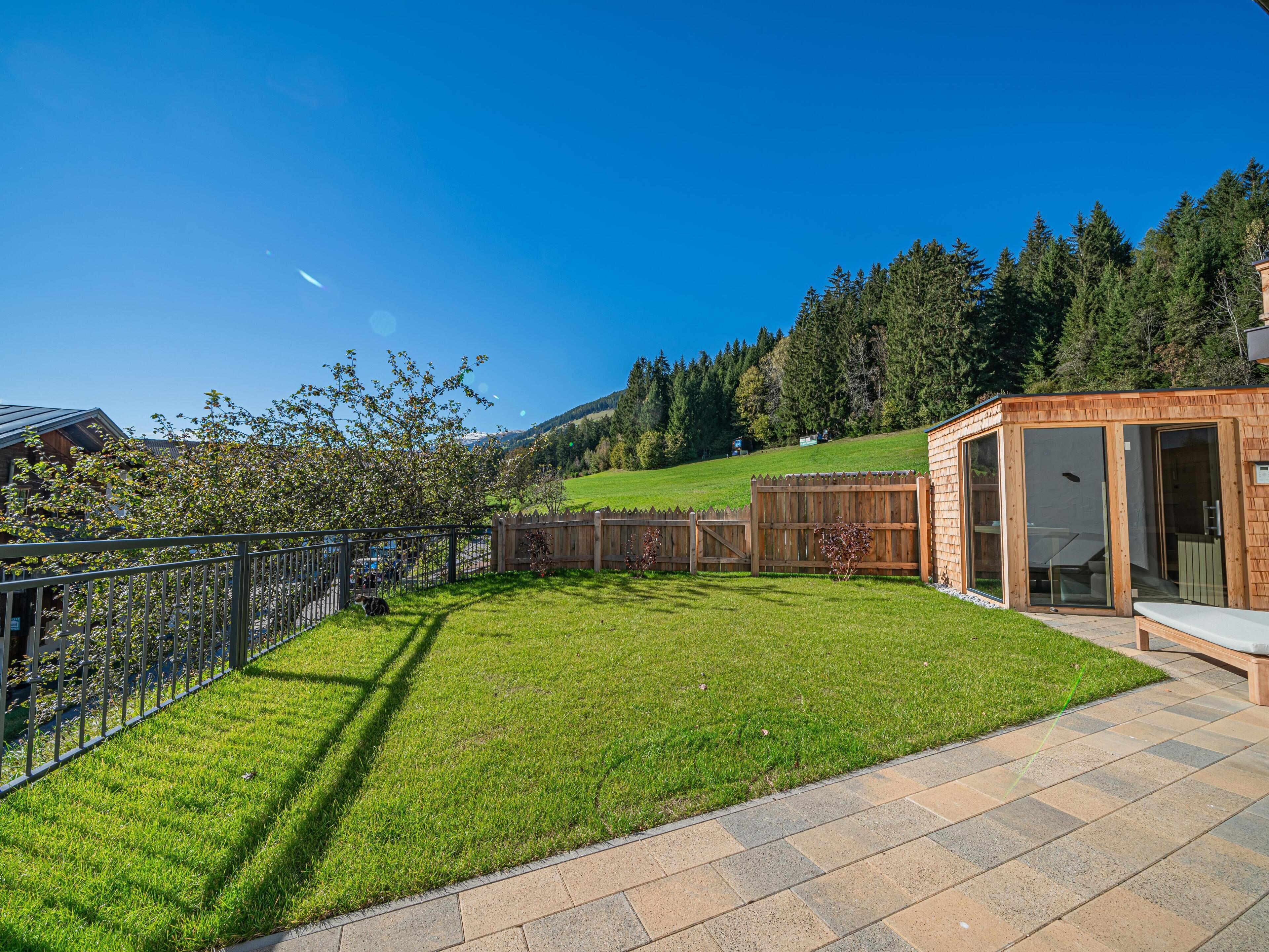 Ein gepflegter Garten mit einer Holzterrasse, einem eingezäunten Bereich und Blick auf eine gebirgige Landschaft.