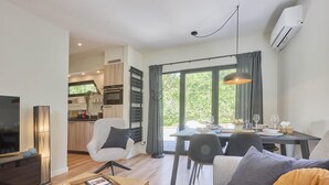 A modern living room with a sofa, a coffee table, and a TV. There is a dining area with a table and chairs. The room has large windows and an air conditioning unit.