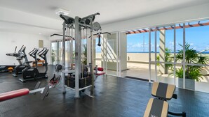 Un gimnasio con máquinas de cardio, equipo de levantamiento de pesas y una banca. Cuenta con ventanas amplias con vista al océano.