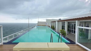 Una piscina con vista al océano, un área de descanso con muebles de mimbre y un edificio con amplias ventanas.