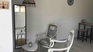 A room with a white-framed armchair, a small round table with a glass container, a hanging light fixture, a wall clock, and a mirror reflecting a dining area. - Maison Basalte studio 1 (Agde)