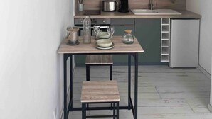 A compact kitchen with a wooden table, a small stool, and a clock on the wall. - Maison Basalte studio 1 (Agde)