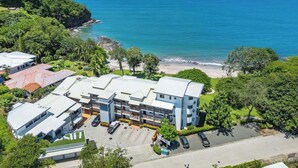 Vista aérea de un hotel costero con varias construcciones, un estacionamiento y una playa al fondo.
