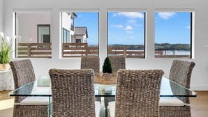 A dining area with wicker chairs and a glass table, large windows with a view of a house and landscape. - Tranquil Tides-Luxury Waterfront home, pool & dock (Kingsland)