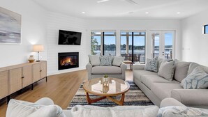 A modern living room with a fireplace, a sofa, a coffee table, and large windows offering a view of the outdoors. - Tranquil Tides-Luxury Waterfront home, pool & dock (Kingsland)