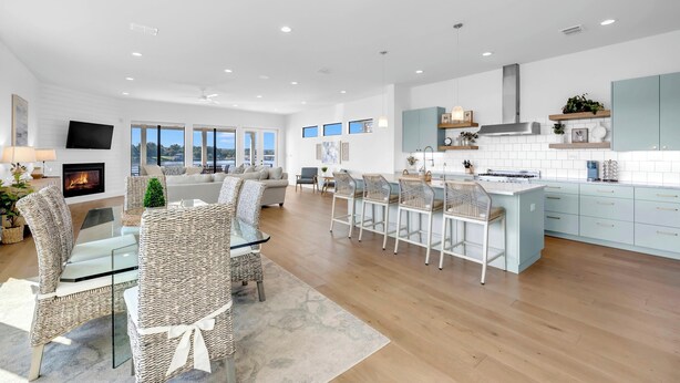A spacious living area with a fireplace, a dining area with wicker chairs, a kitchen with light blue cabinets, and a large window offering a view of the outdoors. - Tranquil Tides-Luxury Waterfront home, pool & dock (Kingsland)