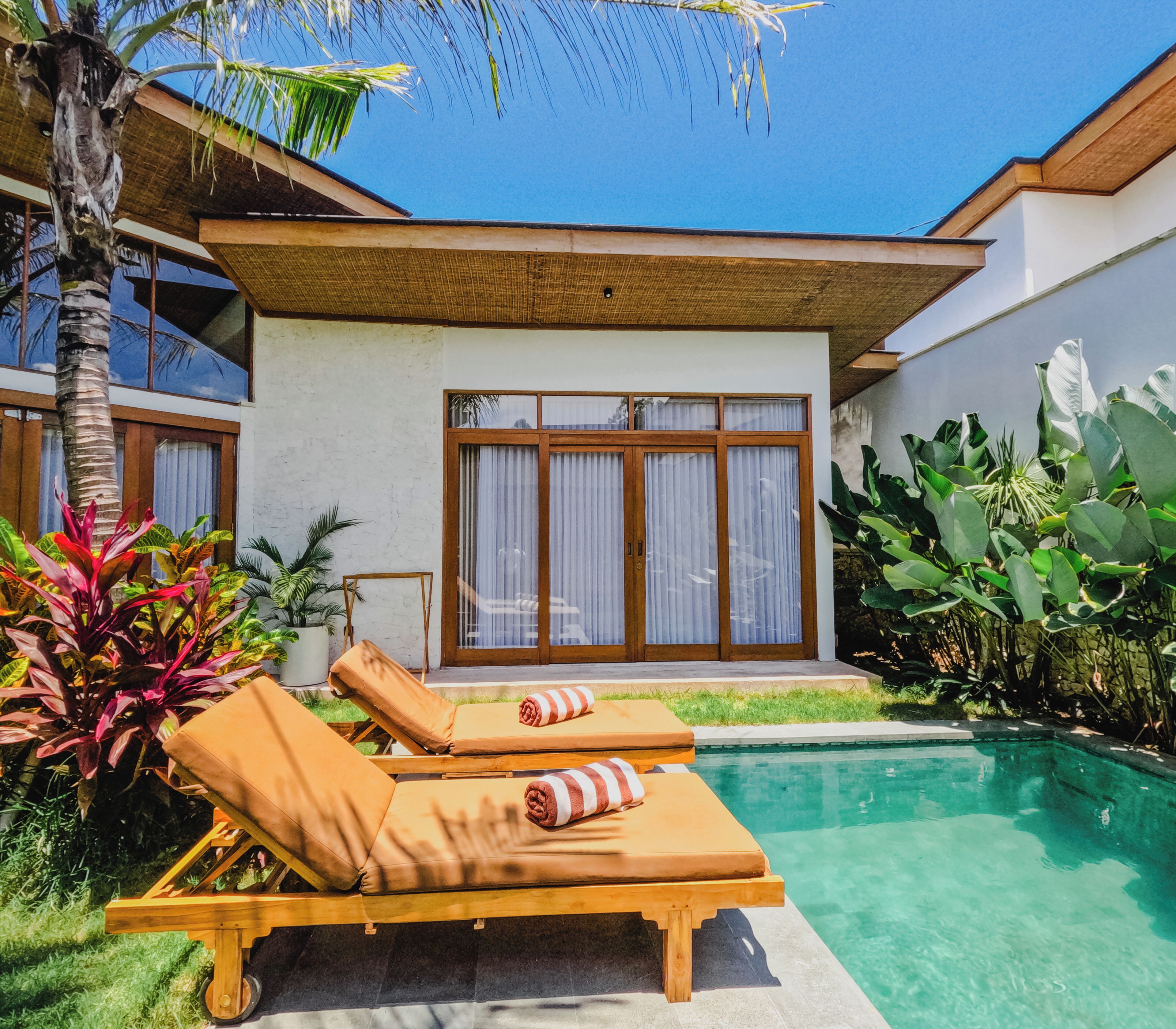 A modern house with a pool, featuring two lounge chairs with towels, a large window, and tropical plants.