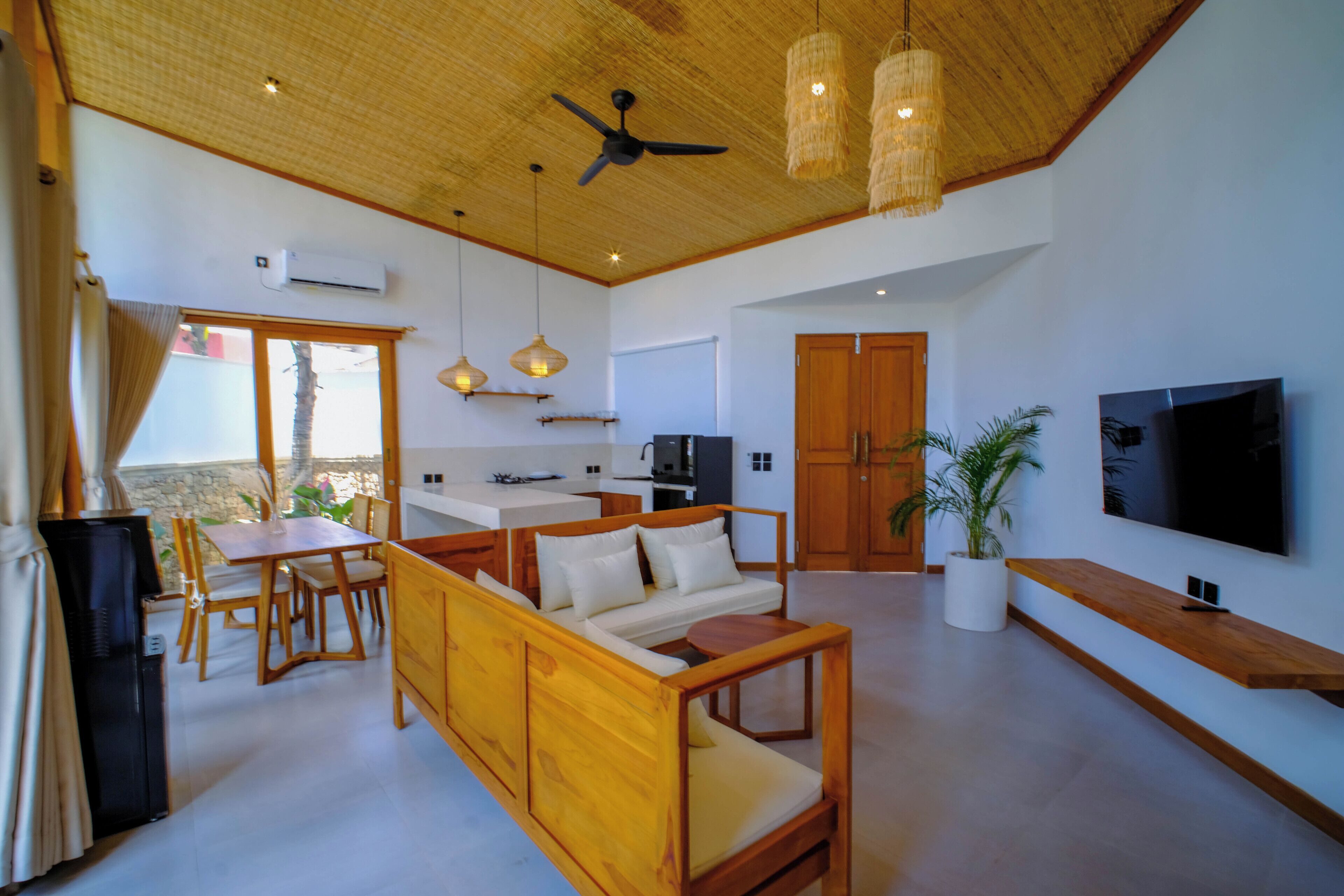 Amplia sala de estar con sofá, mesa de comedor y sillas, zona de cocina, televisor montado en la pared y ventilador de techo.