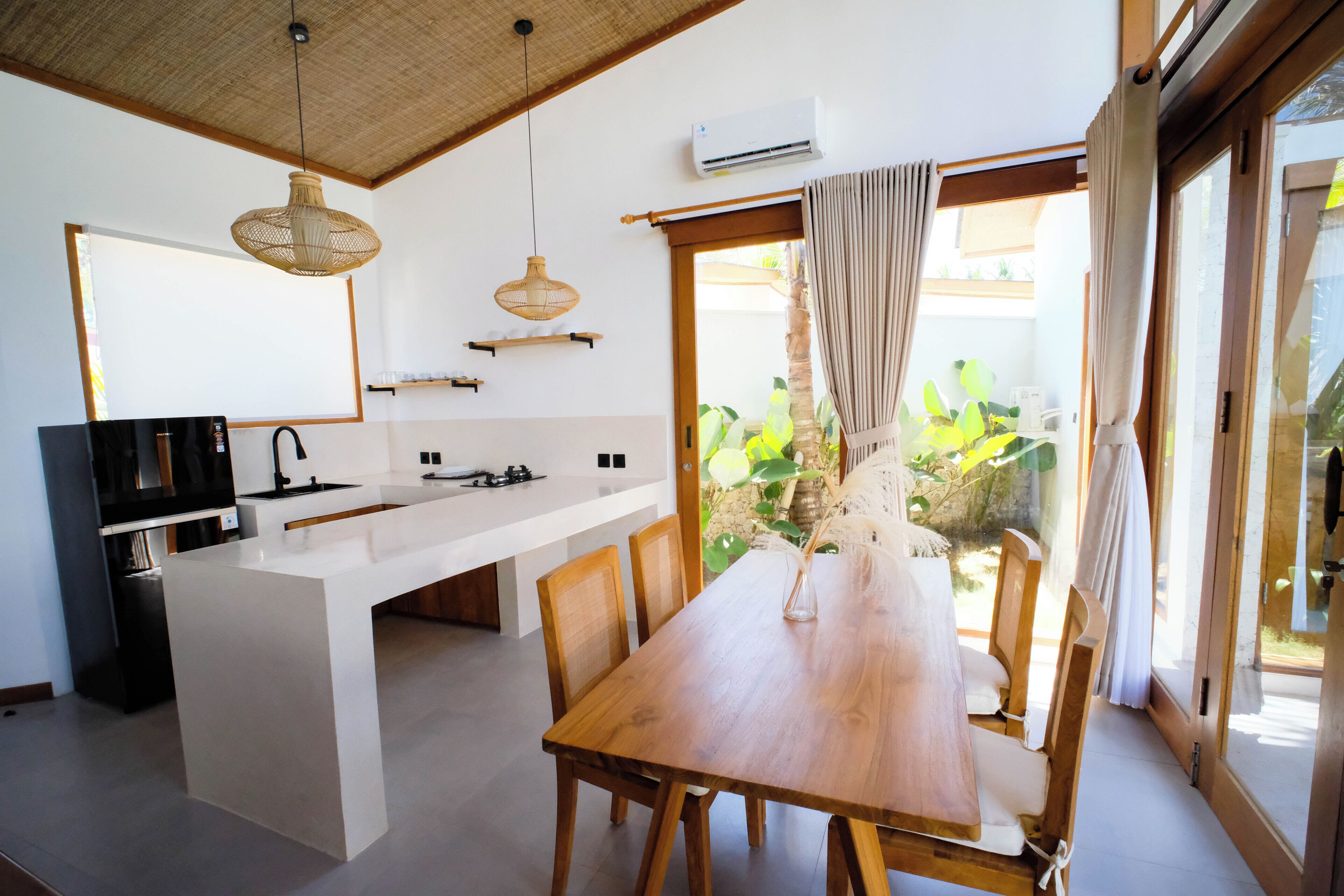A modern kitchen with a central island, wooden dining table, and chairs. There is a large window with curtains, a hanging light fixture, and an air conditioning unit.