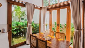 Un comedor con una mesa y sillas de madera, ventanas amplias con cortinas y un equipo de aire acondicionado en la pared.