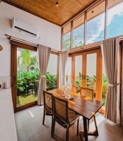 A dining area with a wooden table and chairs, large windows with curtains, and an air conditioning unit on the wall.