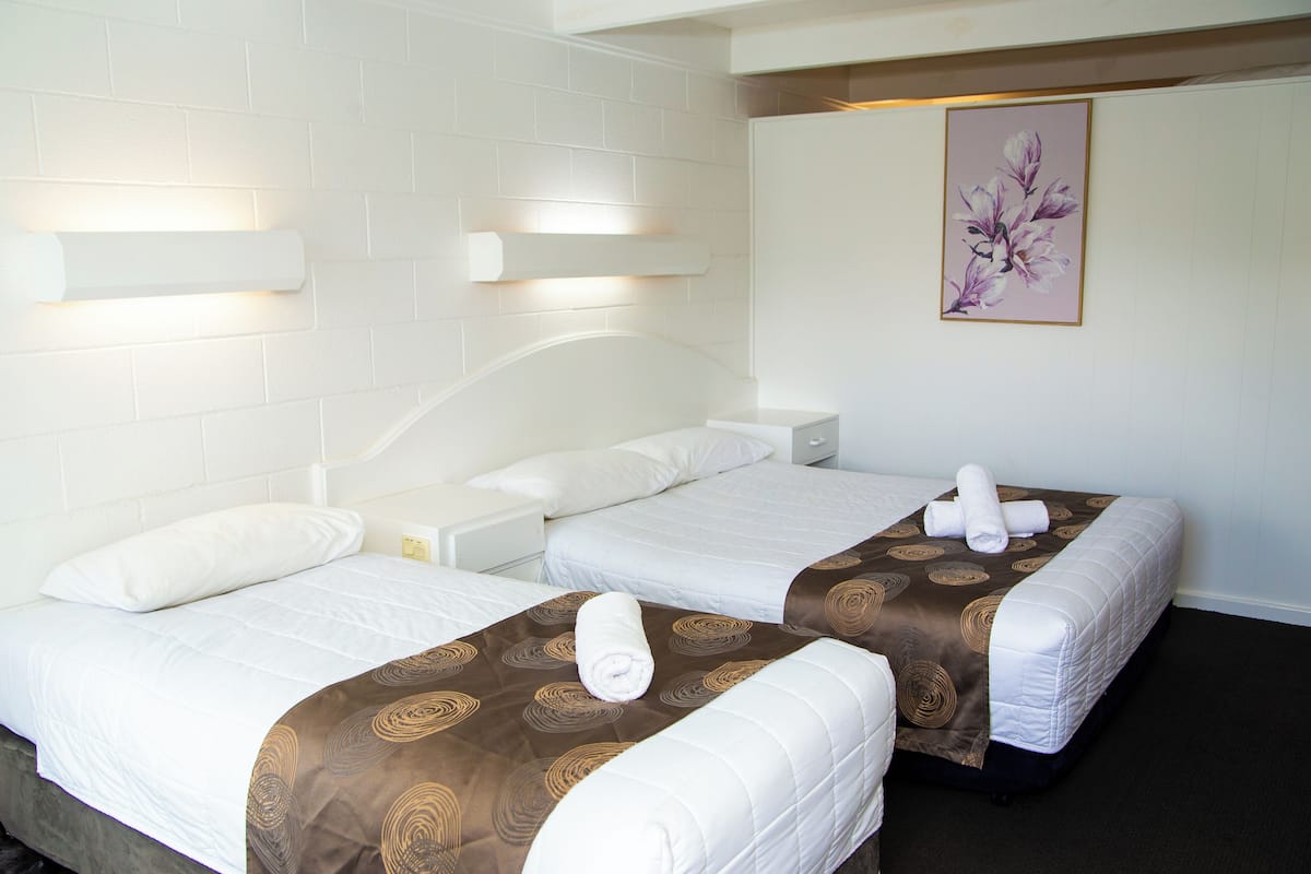 Quarto de hotel com duas camas, roupa de cama branca, colchas marrons estampadas e uma obra de arte floral na parede.