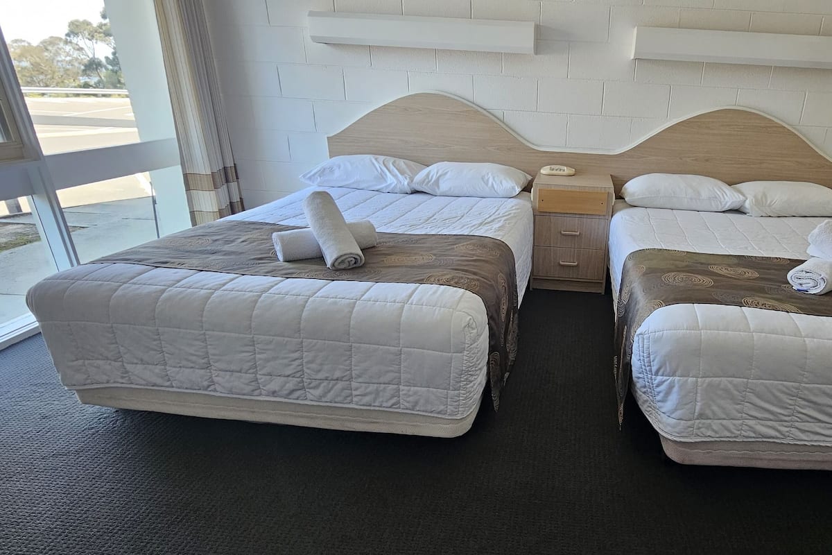 Quarto de hotel com duas camas, cada uma com colchão e almofadas brancas acolchoadas, cabeceira de madeira e pequena mesa de cabeceira com abajur.