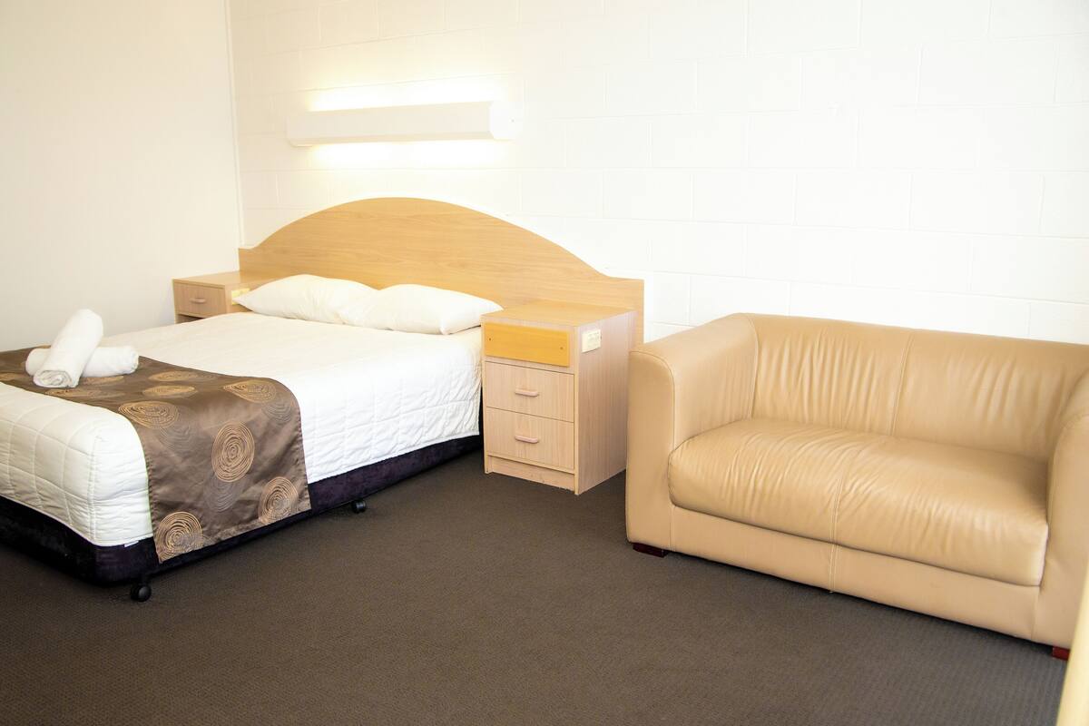 Quarto de hotel com cama, mesa de cabeceira, luminária e sofá bege.