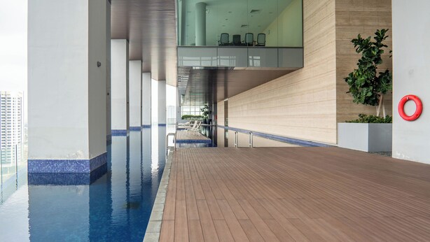 Zona de piscina interior moderna con cubierta de madera, paredes de vidrio y un área de estar con sillas.