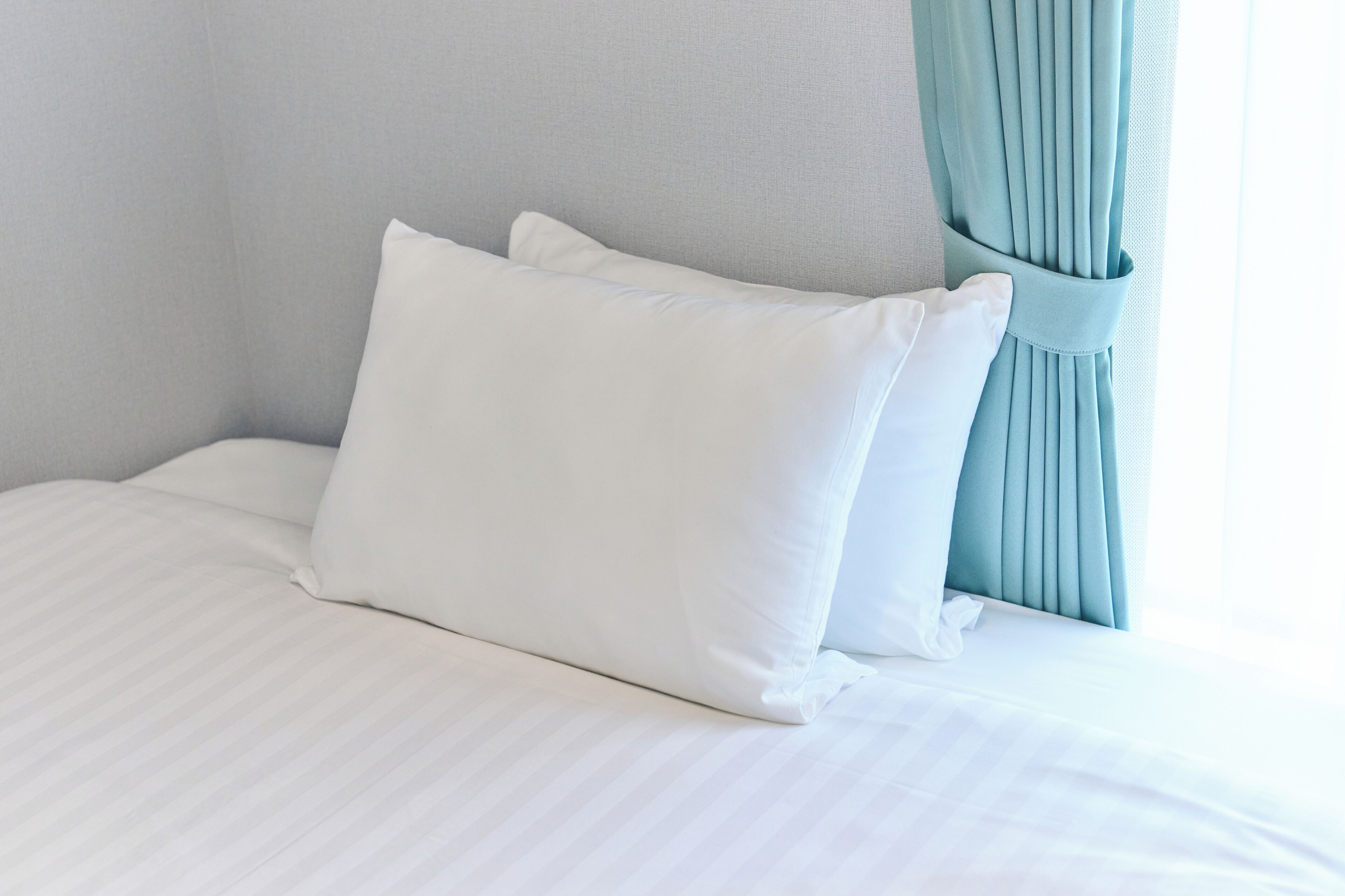 A neatly made bed with white bedding and pillows, a light blue curtain, and a plain wall.