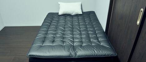 A single bed with a tufted headboard and a white pillow in a room with dark wooden flooring and a dark wooden door.