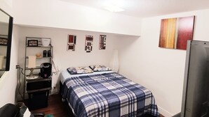A compact bedroom with a bed, a desk, a chair, a TV, and a shelf with personal items.