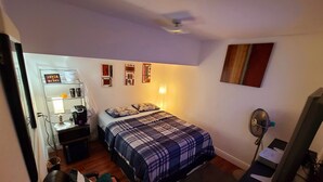 A bedroom with a bed, a desk, a chair, a standing fan, and a wall-mounted shelf with various items.