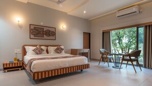 A modern hotel room with a large bed, a desk, and a chair. There is a ceiling fan, an air conditioning unit, and a window with curtains. - Kings Kraft River Side Resort  (Talala)