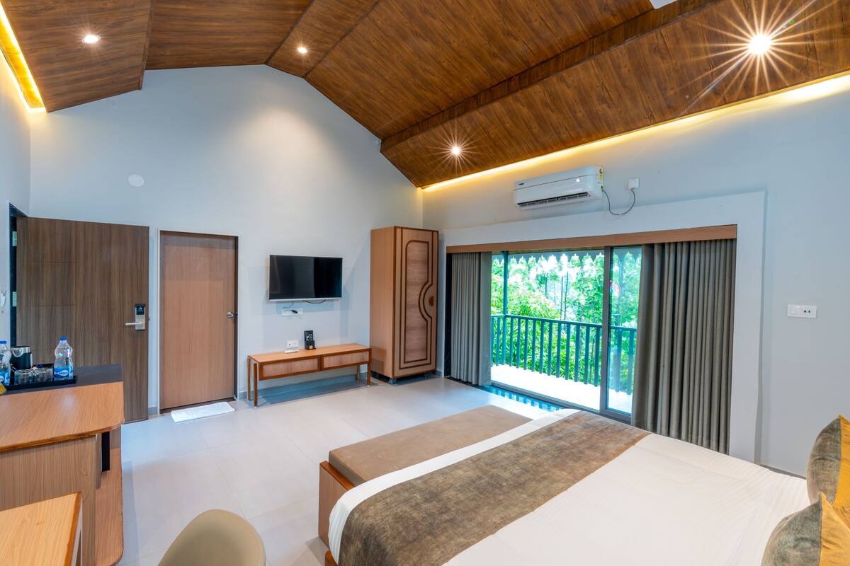 A modern hotel room with a large bed, a desk, a TV, and a balcony with a view of greenery.