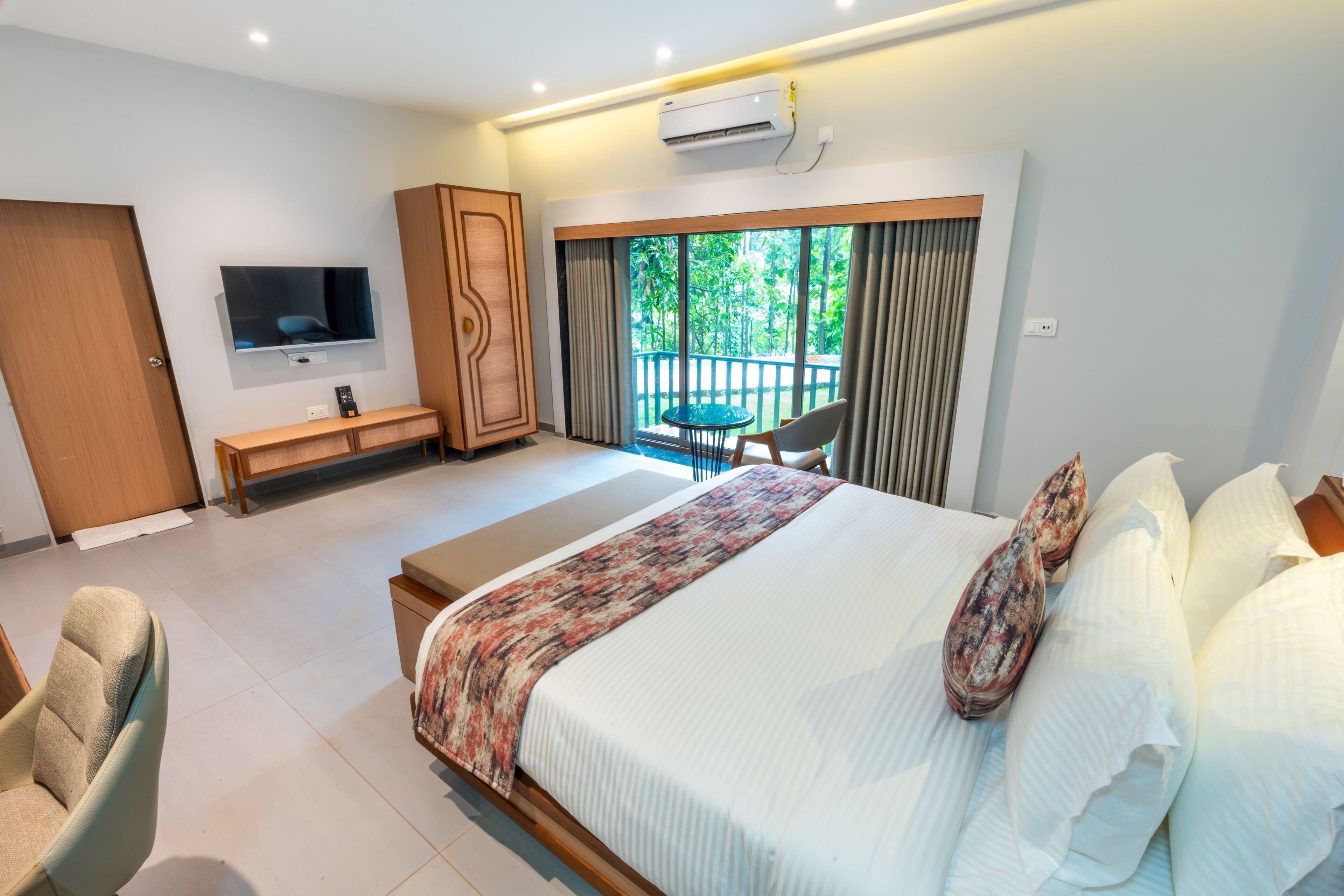 Ein modernes Hotelzimmer mit einem großen Bett, einem an der Wand montierten Fernseher, einem Schreibtisch, einem Stuhl und einem Balkon mit Blick auf Grünflächen.
